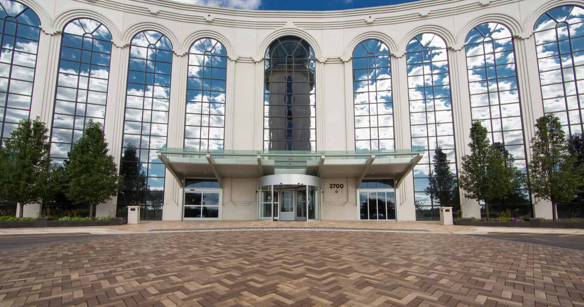 A modern, curved building with a grand entrance. Tall, arched windows reflect a blue sky with white clouds. The entrance features a glass canopy, a revolving door, and sliding doors, with the number 2700 visible. The foreground is paved with herringbone brickwork.
