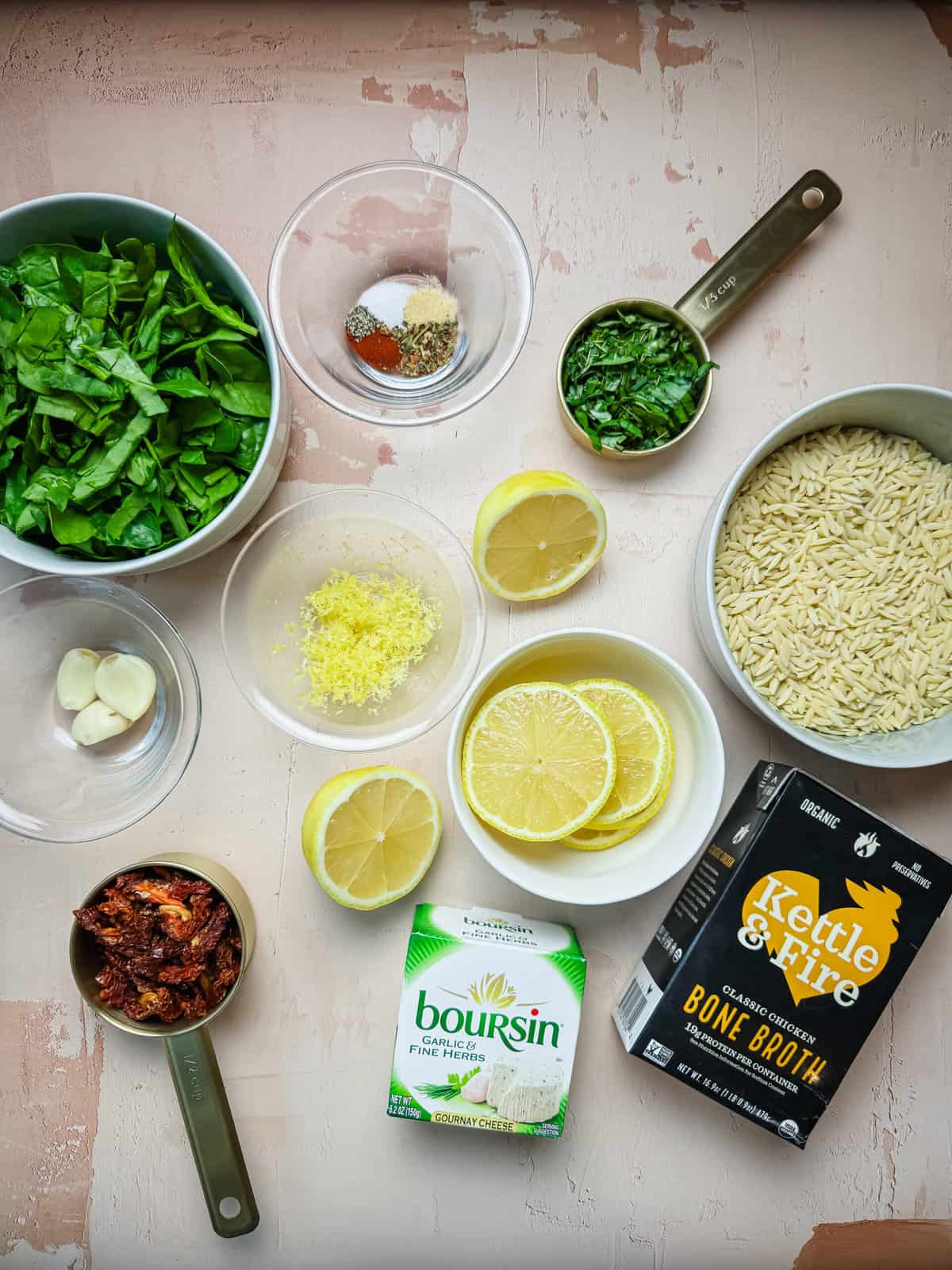 All of the ingredients that go into this dish pictured from a top view. Spinach, seasonings, fresh herbs, orzo, lemon, garlic, lemon zest, sundried tomatoes, boursin, chicken bone broth