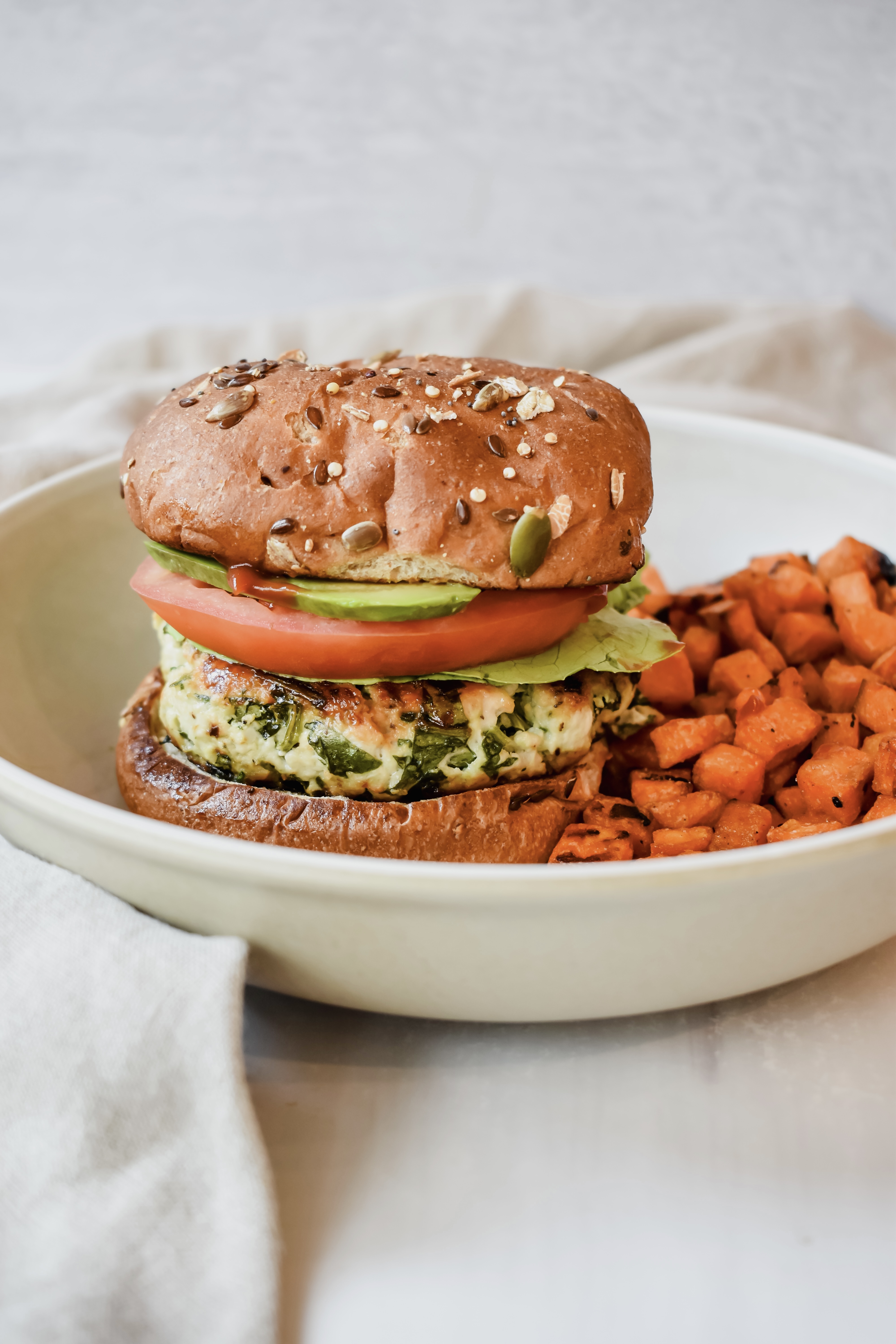 This is a side view of my chicken feta spinach burgers on a whole grain bun with tomatoes, lettuce, avocado and ketchup served with a side of roasted sweet potatoes. 