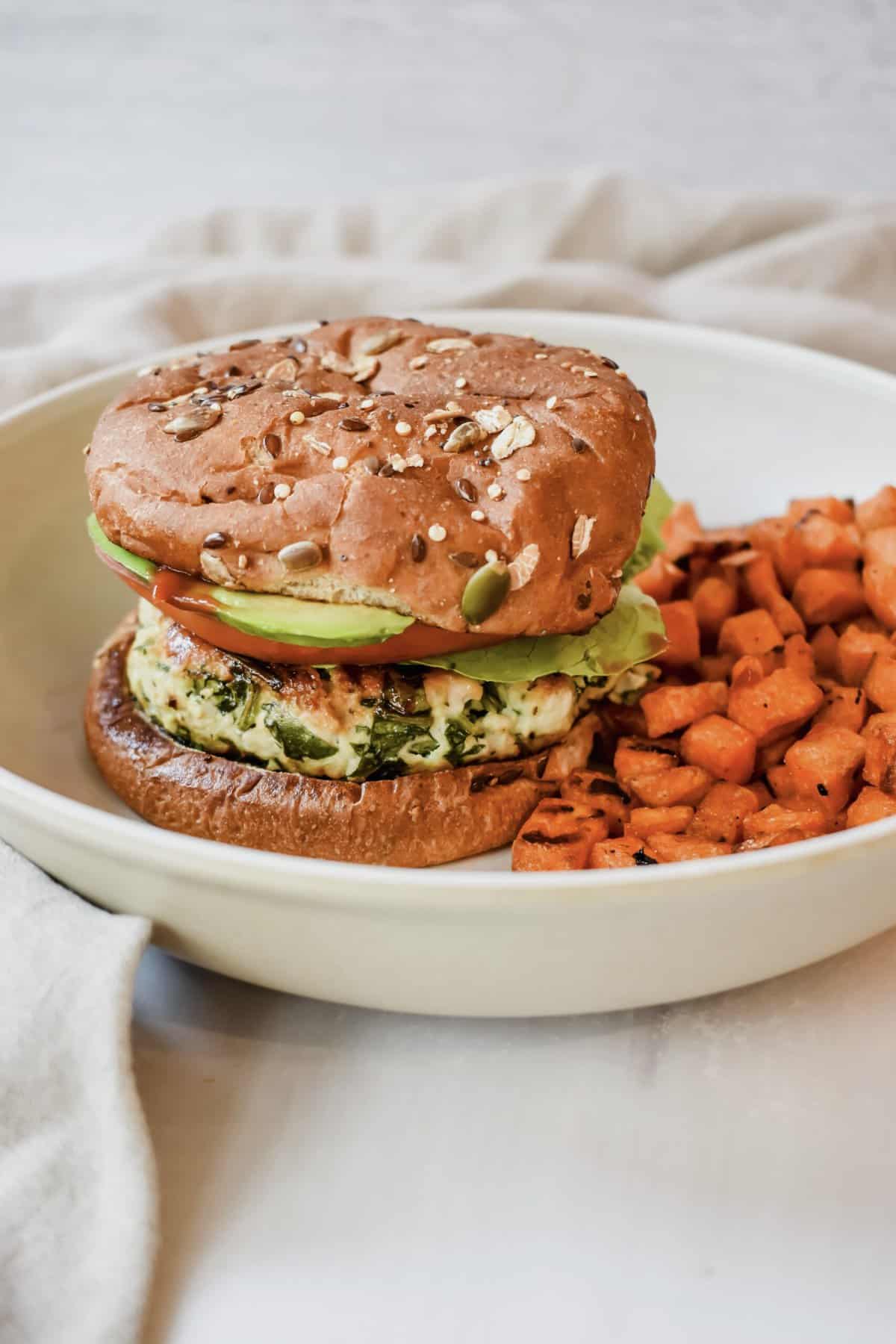 This is a close up side view of my chicken feta spinach burgers on a whole grain bun with tomatoes, lettuce, avocado and ketchup served with a side of roasted sweet potatoes. 