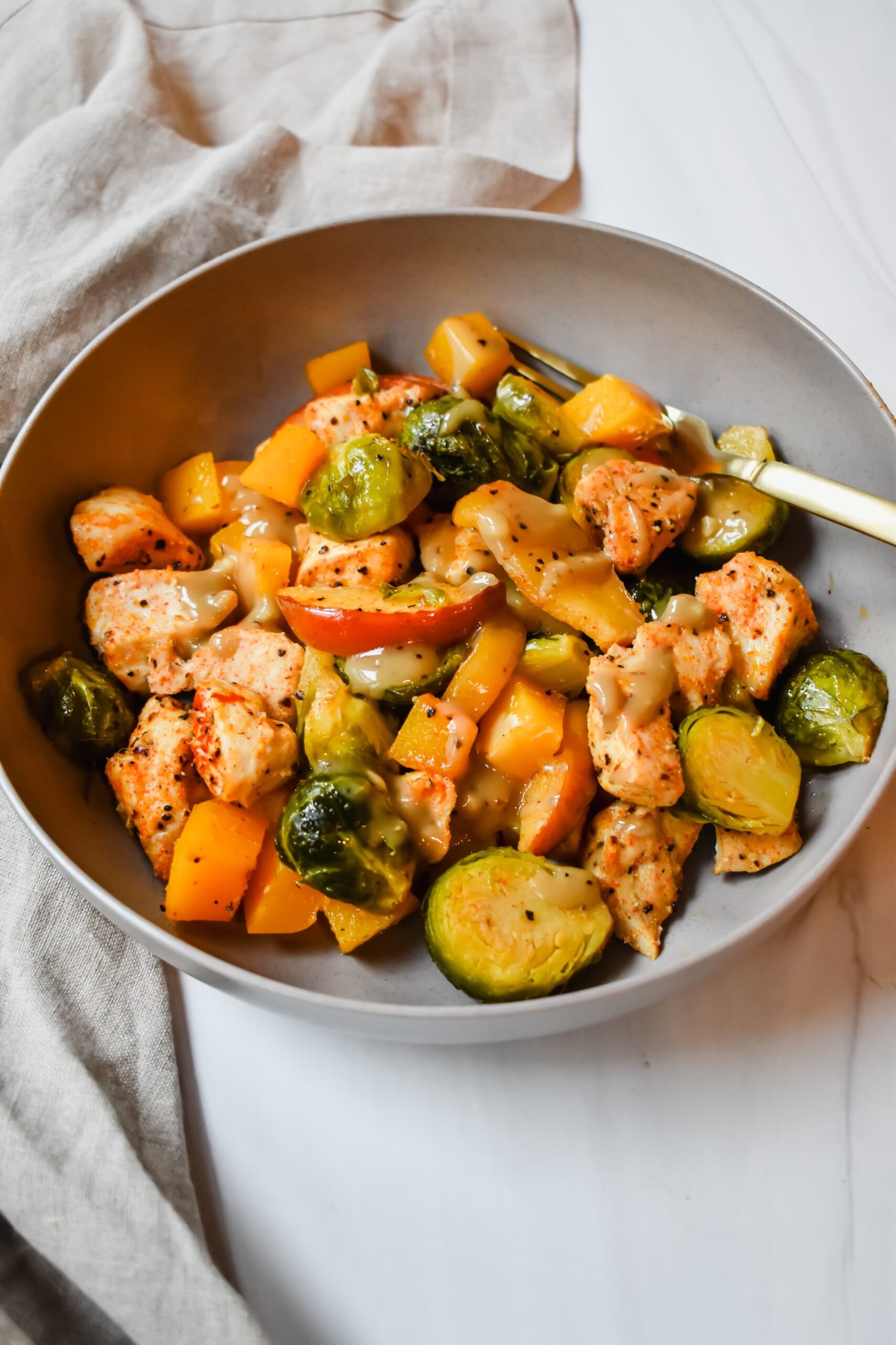 This is a side view close up of my fall sheet pan chicken and veggies dish which is topped with a honey tahini glaze and in a bowl with a gold fork. 