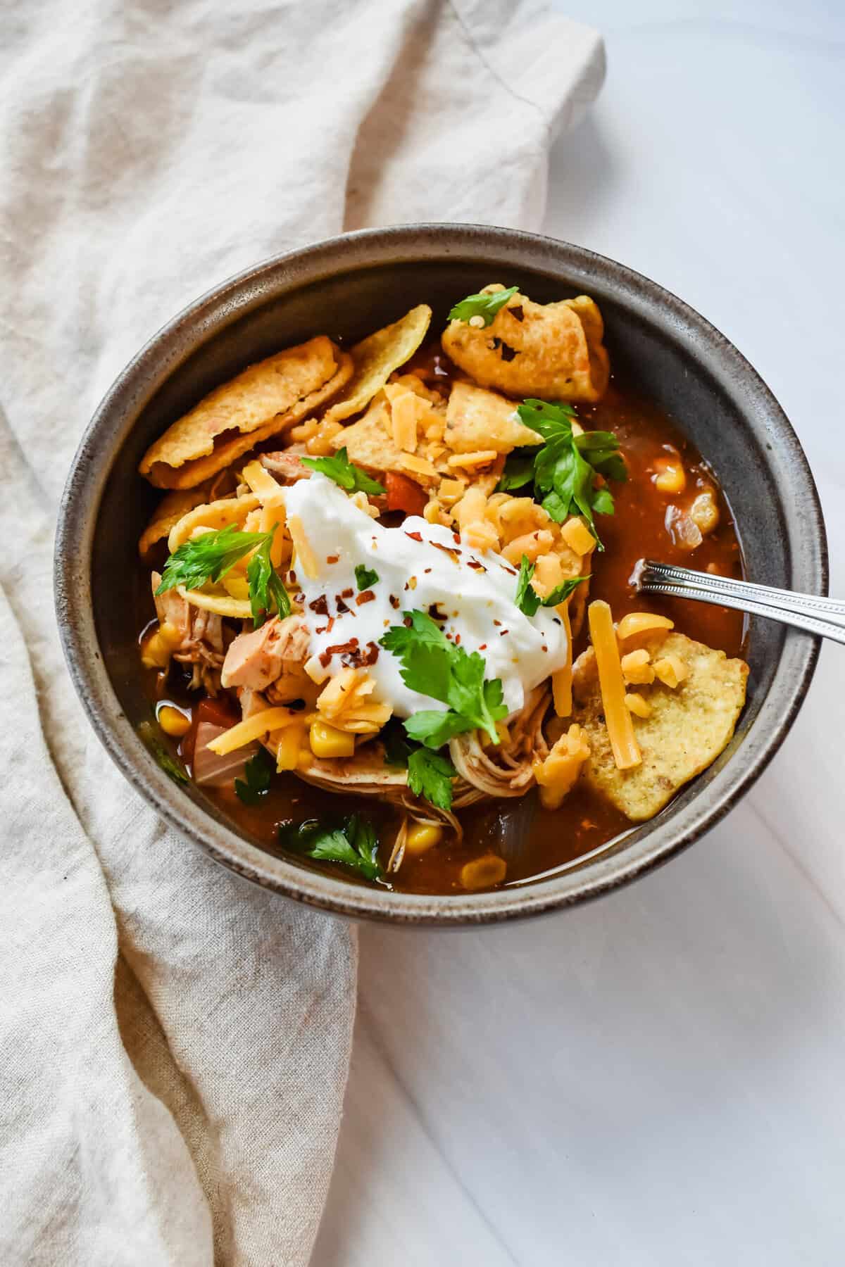 This is a side view of my crockpot southwest chicken soup in a bowl topped with corn chips, greek yogurt, mexican cheese and red pepper chili flakes.