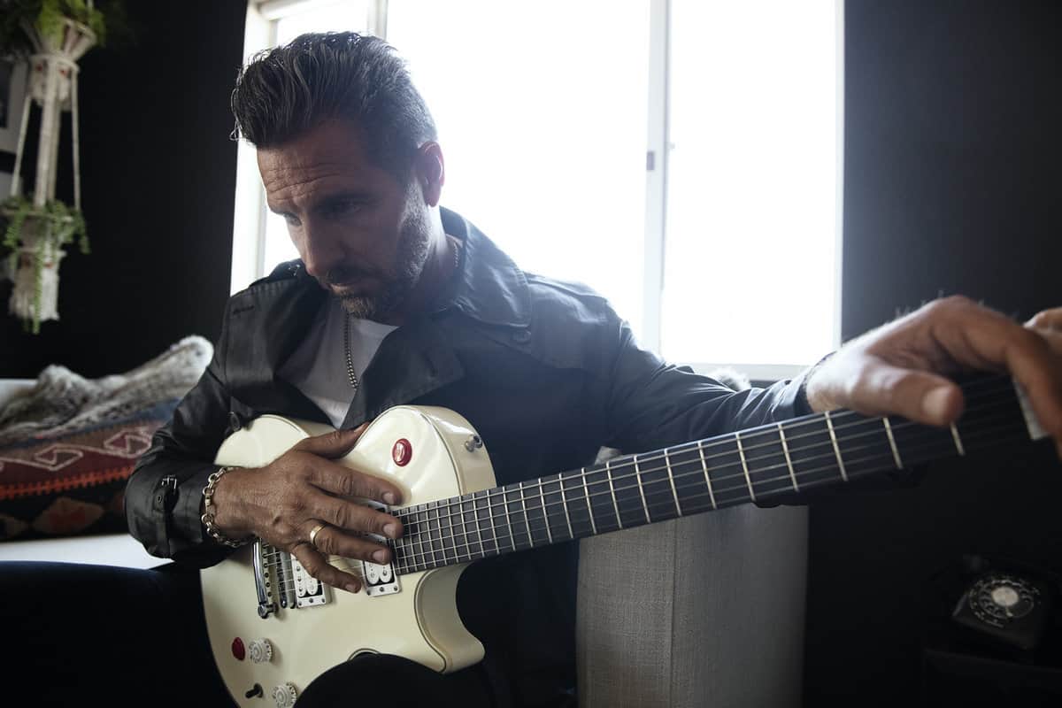 Ed Quinn holding his guitar sitting in front of a bright window deep in thought as if developing lyrics for new music