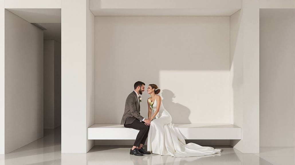 Elegant wedding couple seated on a modern white bench in a minimalist setting.