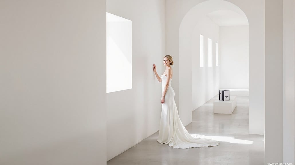 Bride in elegant wedding gown standing in bright, minimalist wedding venue with white walls and natu.