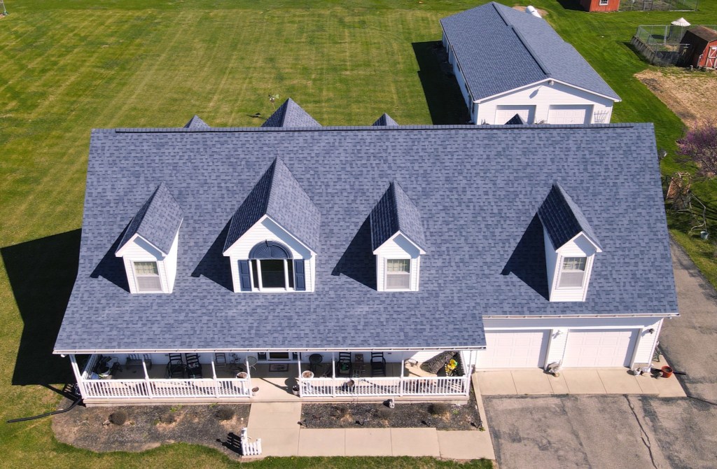 Aerial view of a house with a new blue asphalt shingle roof, featuring multiple dormer windows, surrounded by a green lawn and a spacious driveway.