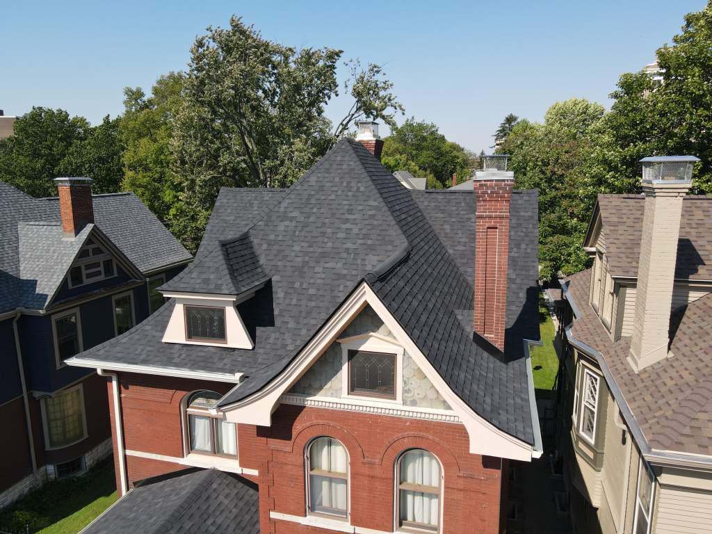 Vivid aerial view of Victorian-style house with new dark asphalt shingle roof, surrounded by lush green trees and neighboring homes, showcasing high-quality roofing and siding.
