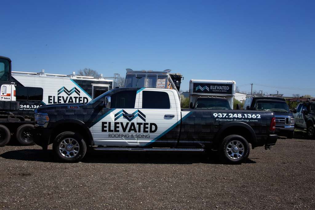 Bold black and white Elevated Roofing and Siding company truck parked on gravel lot with other service vehicles in the background, showcasing professional roofing and siding team.