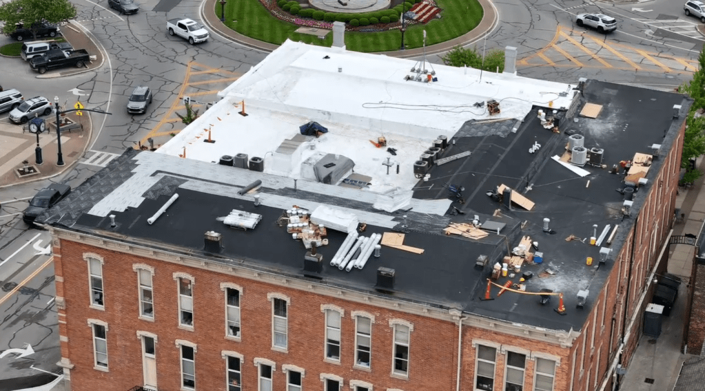 New roof installation with black and white roofing materials on brick commercial building. Roofing tools, materials, and construction debris visible on flat rooftop during ongoing roof replacement.