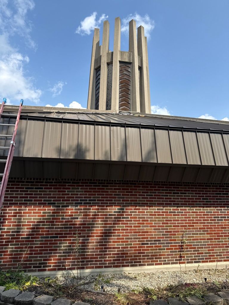 Bright blue sky with clouds, showing the roof and steeple of a church or historic building with a brick wall and metal roofing in the foreground.