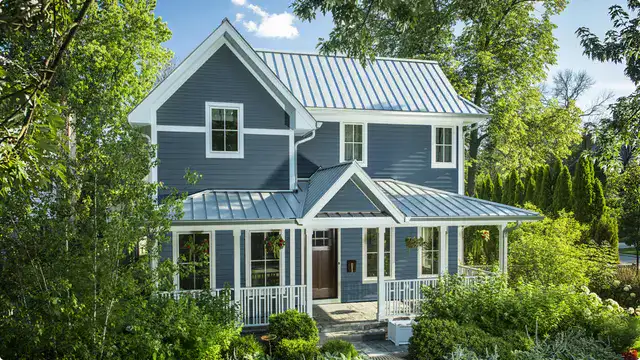 Blue siding home with metal roof, lush green landscaping, and white accents, showcasing residential renovation, roofing, and siding experts.