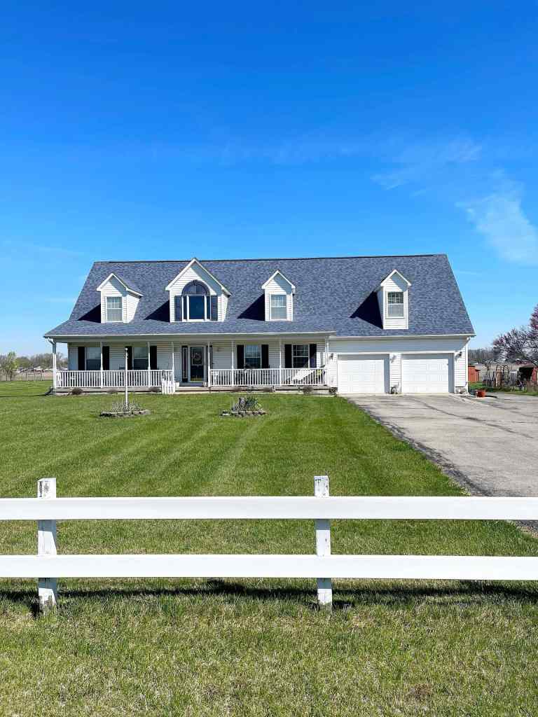 A white house with dormer windows and a wraparound front porch featuring a blue shingle roof and a white fence.