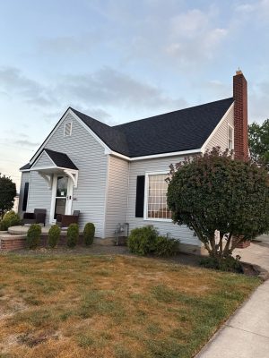 Enhanced residential home with new black asphalt shingle roof, white siding, black shutters, and brick chimney, showcasing professional roofing and siding installation services.