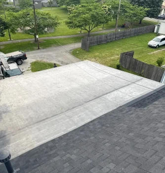 Aerial view of a residential roof showing completed flat roofing installation with a gray surface, surrounded by trees, fences, and neighboring houses, highlighting expert roofing services.