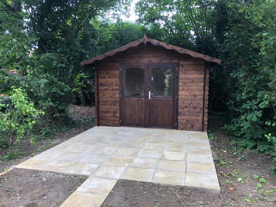 Wooden garden shed with tiled patio in lush green garden setting.