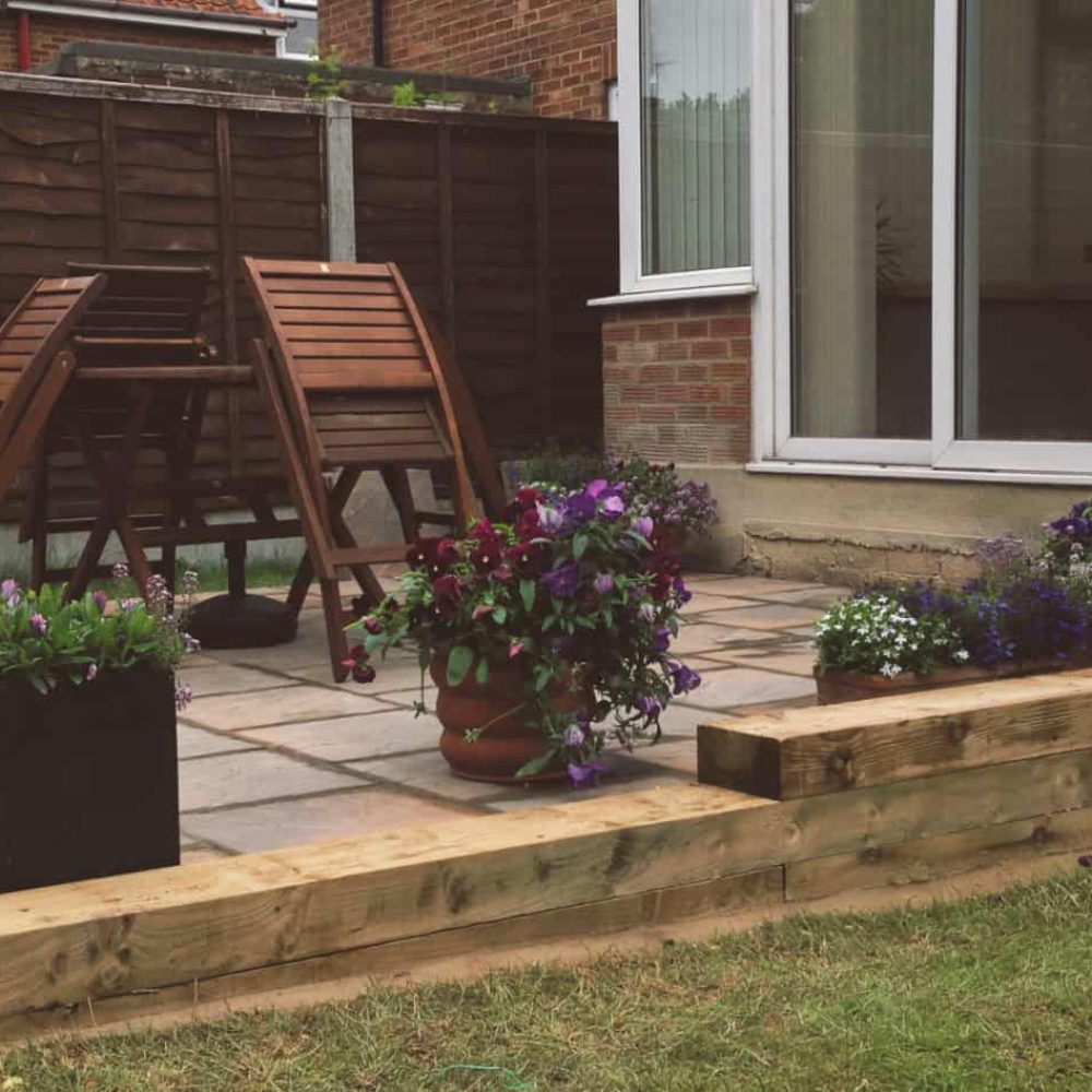 Outdoor garden patio with wooden table and chairs, potted plants, and brick house background.