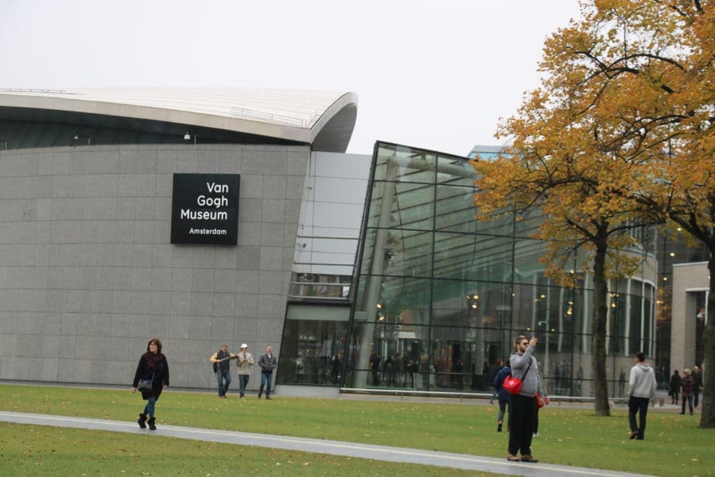 A grey curved building with a sign reading "Van Gogh Museum". People are walking to the entrance of the building over a grassy lawn.