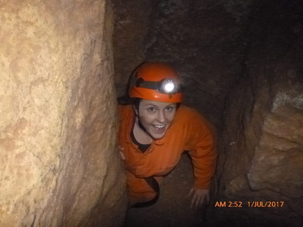 Emma is crawling through a cave dressed in an orange jumpsuit with a hard hat and torch