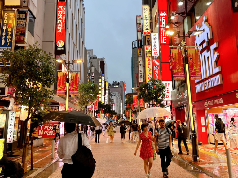 Tokyo city street at dusk with bright neon signs and pedestrians with umbrellas, capturing vibrant urban nightlife in Japan.