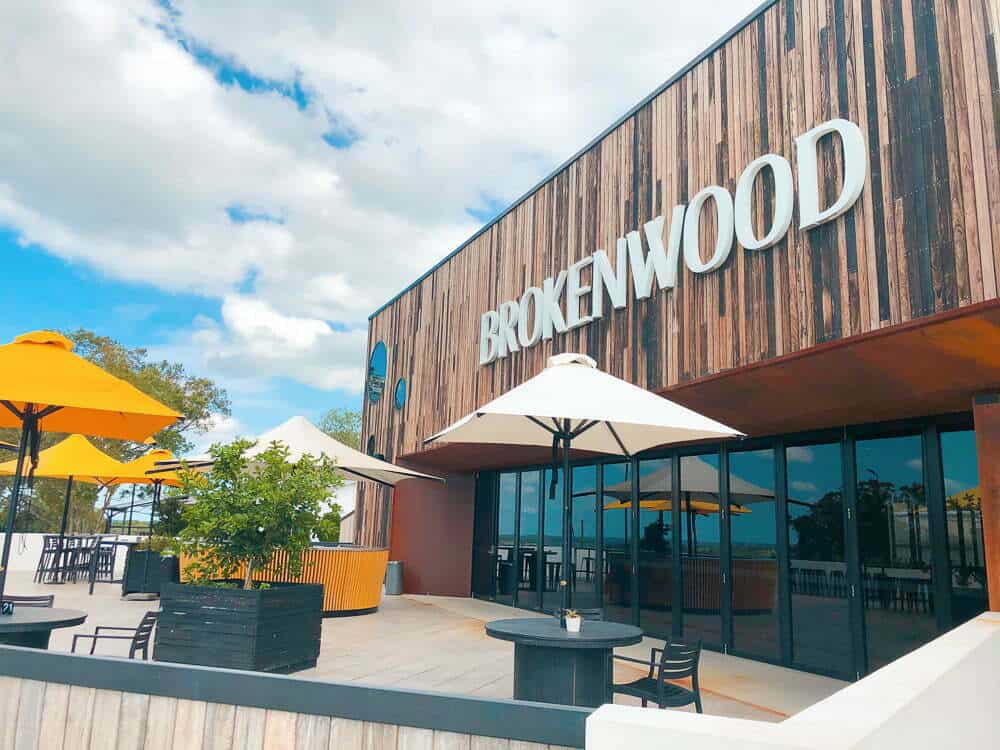Modern shopping centre exterior with "Brokenwood" signage, outdoor seating with yellow and white umbrellas, lush greenery, and a clear blue sky.