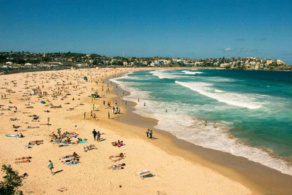 Golden beach with sunbathers and swimmers enjoying the sunny weather, with a scenic coastal town in the background. Ideal for seaside holidays, beach activities, surfing, relaxing, and family vacations along the Australian coast.