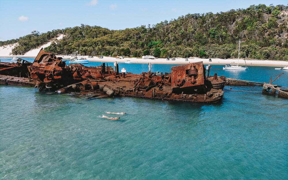 Rusty shipwreck on a peaceful beach with boats and lush green hills in the background in Australia.
