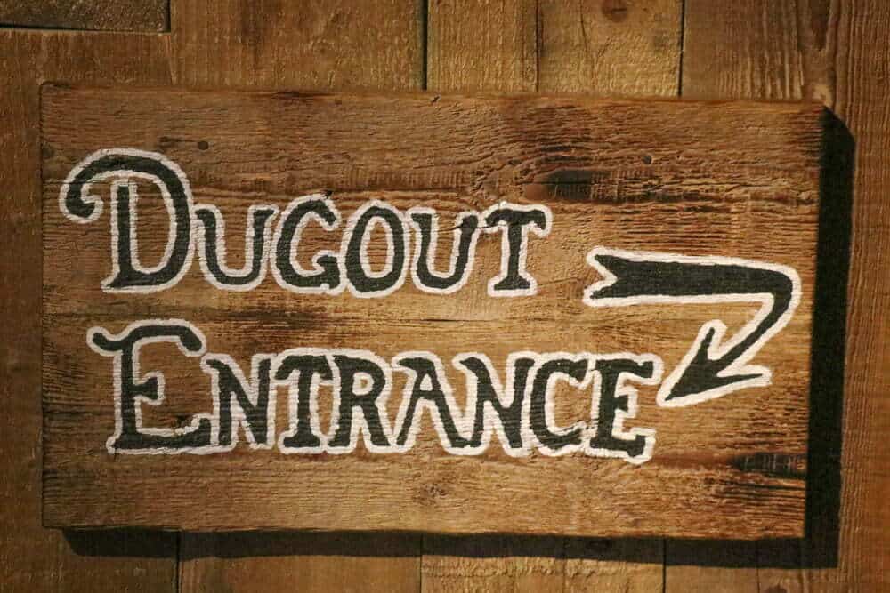 Rustic wooden sign with "DUGOUT ENTRANCE" written in chalk-style font and an arrow indicating direction, suitable for sports venues or baseball fields.