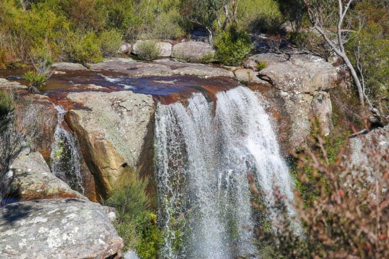 Waterfall in lush Australian bushland with flowing water over rocks, surrounded by native trees and greenery, ideal for hiking and nature photography.