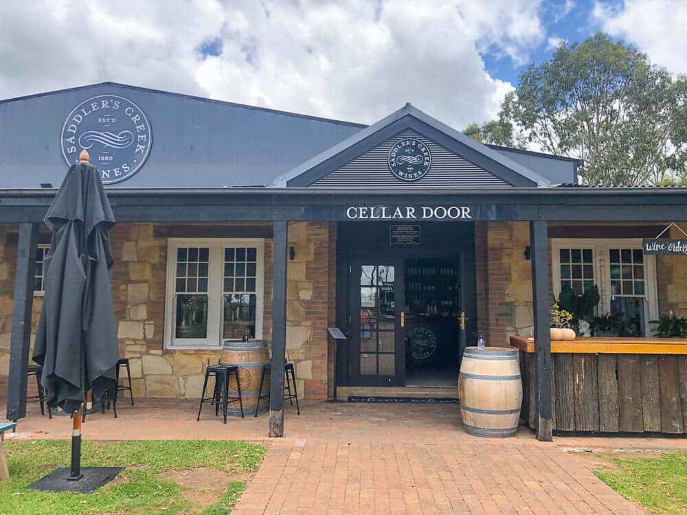 Rustic winery entrance at Saddler’s Creek Wines in Hunter Valley, featuring a cellar door with outdoor seating, wine barrels, and charming stone and wood details, under a blue sky with trees in the background.