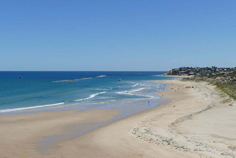 Soft sandy beach with calm blue waters and a distant pier, perfect for relaxation and seaside adventures along Australia's stunning coastline.