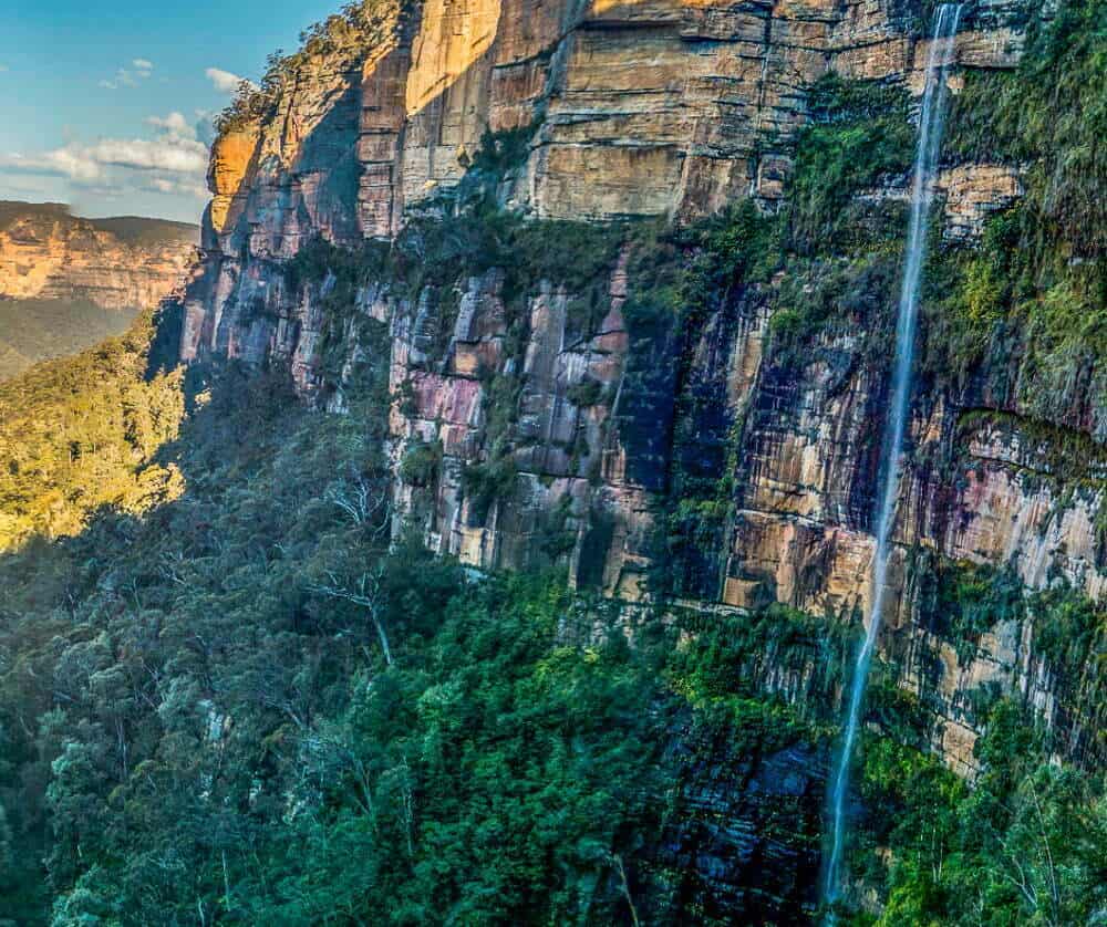 Vivid image of a cascading waterfall flowing down a rugged cliff face surrounded by lush green forest in a scenic natural landscape, perfect for adventure and nature tourism enthusiasts.