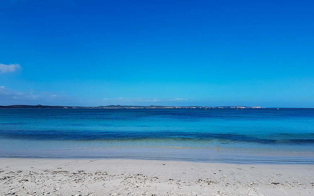 Aerial view of a pristine beach with white sand and clear turquoise waters, under a bright blue sky with minimal clouds, perfect for relaxing coastal adventures and seaside exploration.