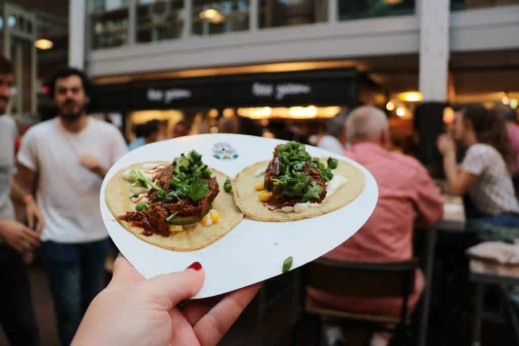 My hand holding up a plate of two beef tacos with Foodhallen patrons in the background