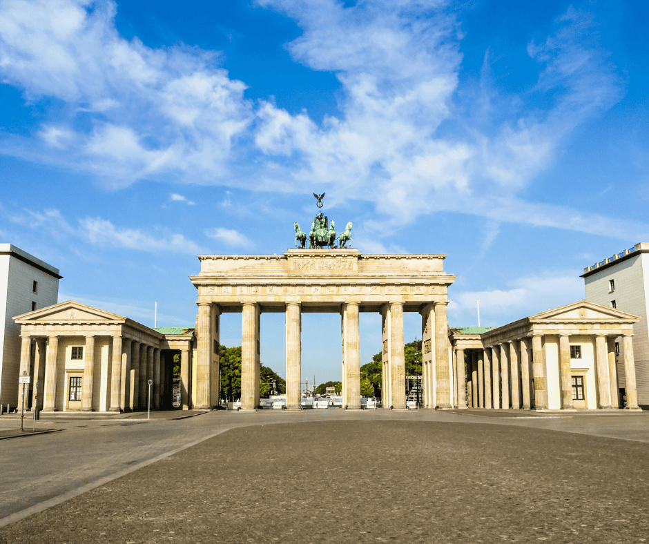 The Brandenburg Gate stands at the end of a paved road. It has 6 large columns and on top is a green statue of horses pulling a chariot.