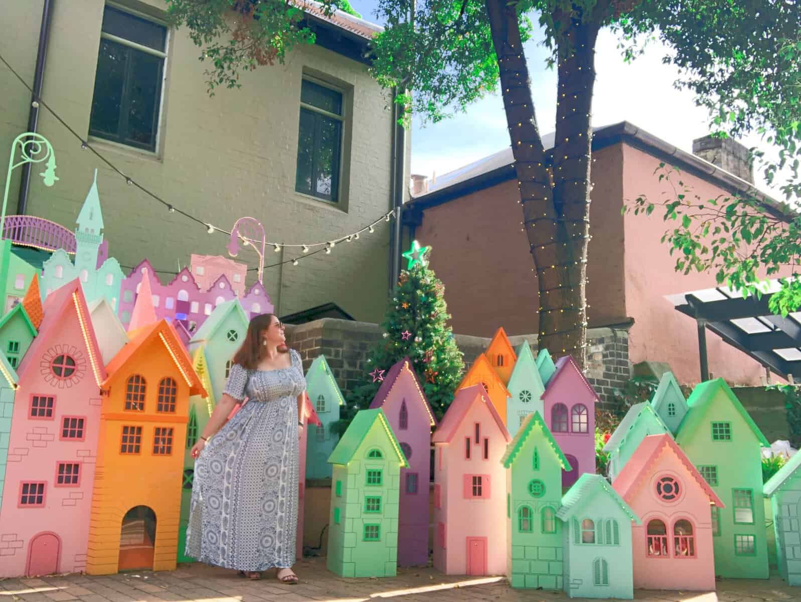 Emma is standing in front of a collection of miniature coloured houses. She is staring to her left at the sky.