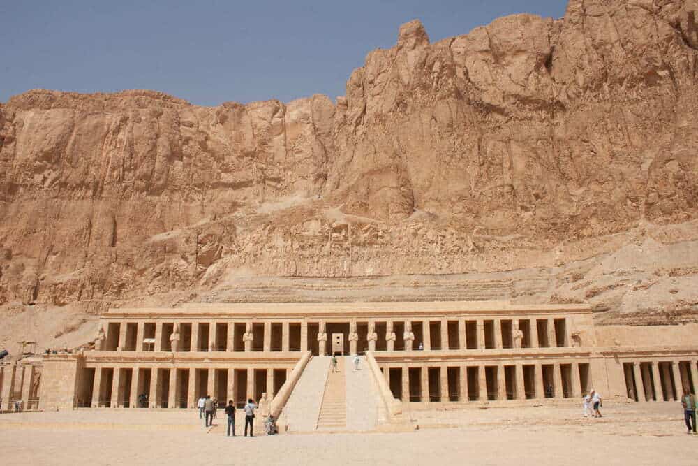 A large temple complex carved into the sandy rock. A cliff rises behind the temple.