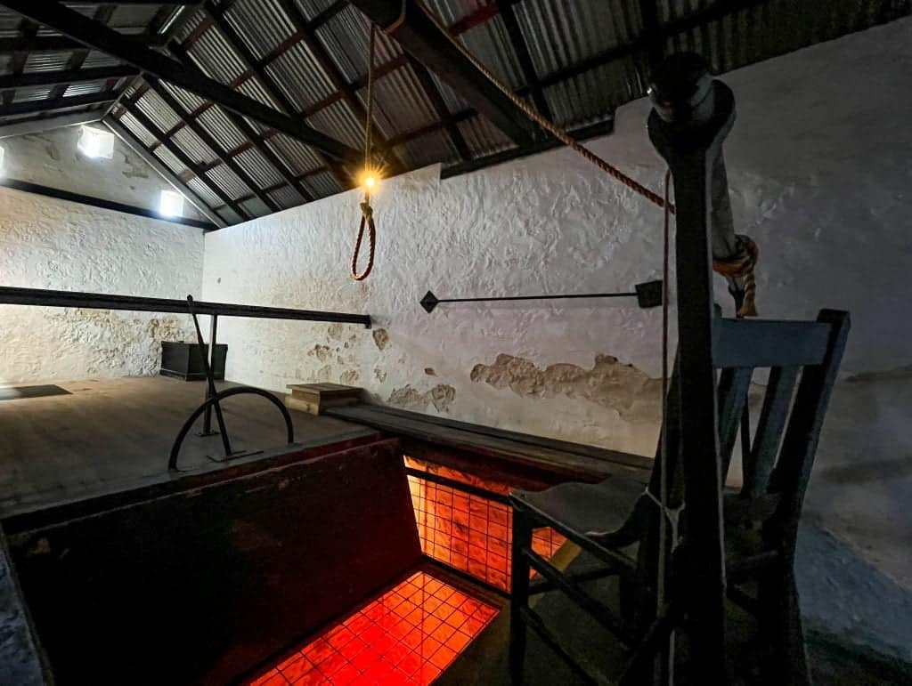 A hangman's noose hangs from the gallows over an open trapdoor illuminated in an eerie red light.