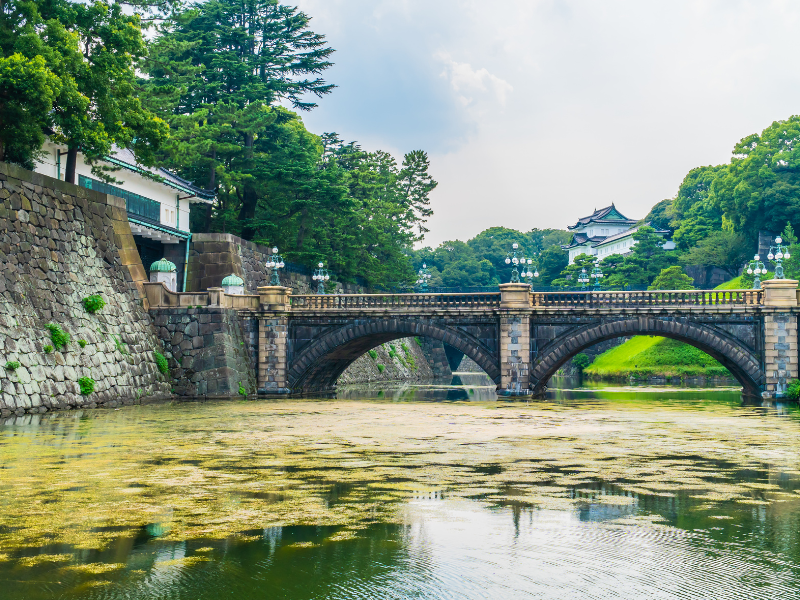 Historic Japanese bridge with lush greenery and traditional architecture on the riverbank.