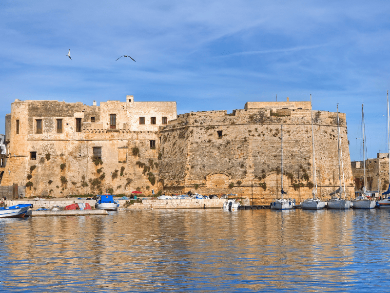 Ancient fortress and marina on the coast of Malta with historic stone walls and sailboats under a blue sky.