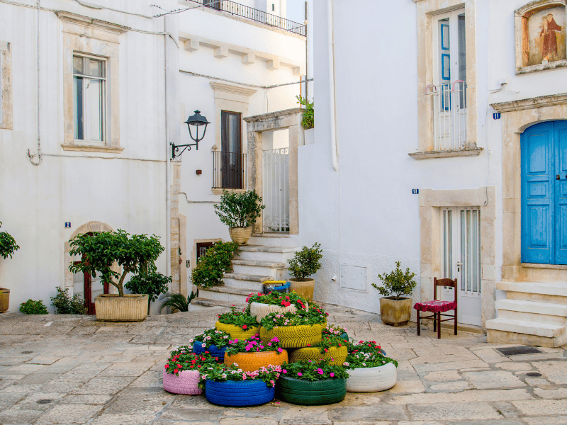 Bright colourful flower arrangements in painted tires on a quaint European cobblestone street with whitewashed buildings, characteristic of Mediterranean travel destinations in Southern Europe.