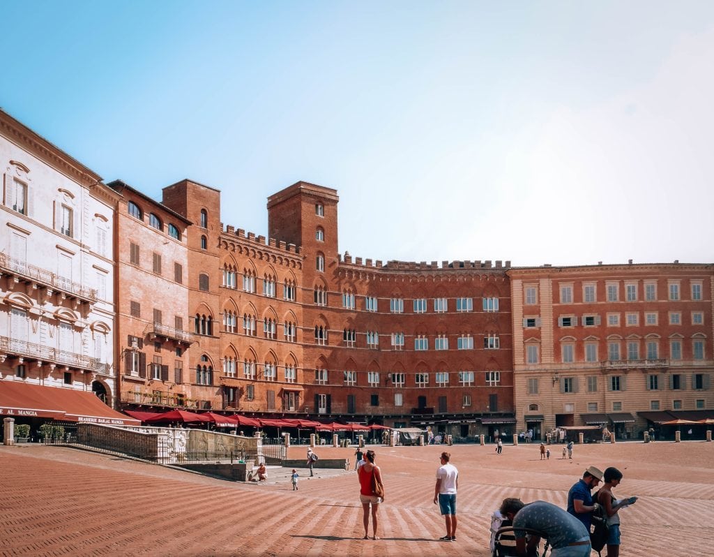 Vibrant red and beige historic buildings in an open square with shops and cafes, capturing authentic European architecture and lively city atmosphere in Italy.