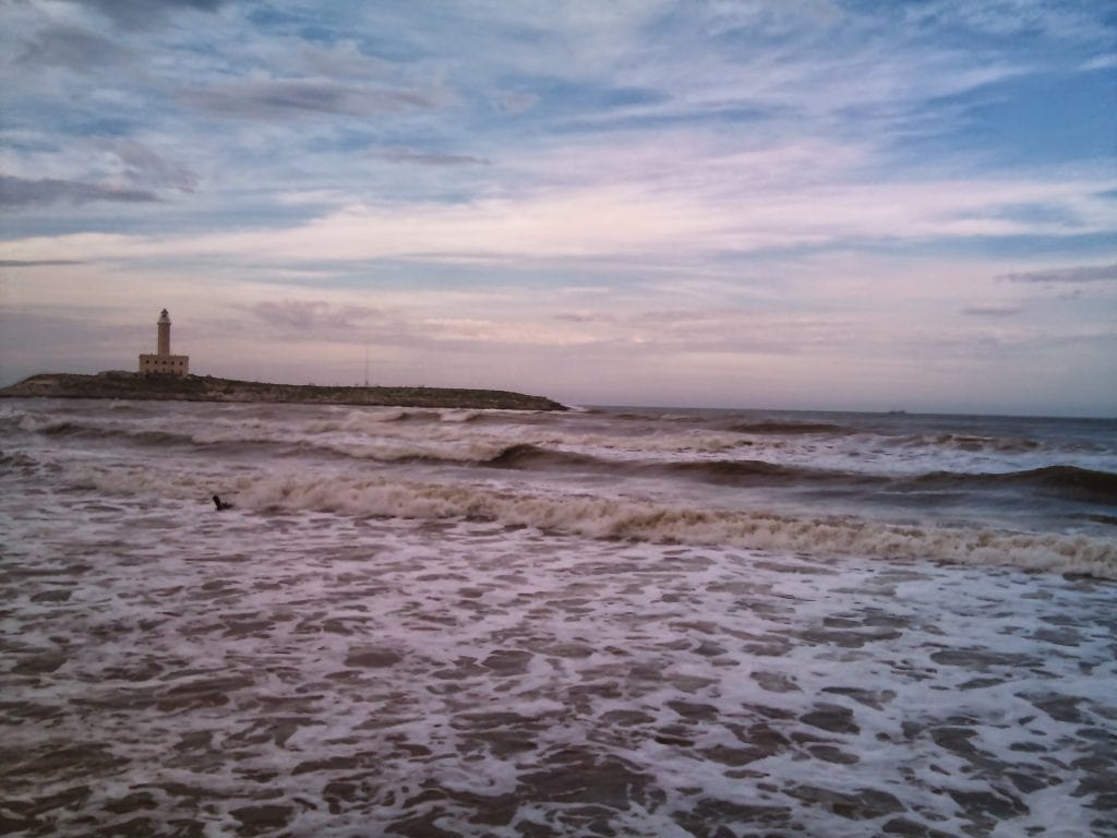 Sea waves crashing on the shore with a lighthouse in the distance at sunset, showcasing coastal scenery and travel adventures.