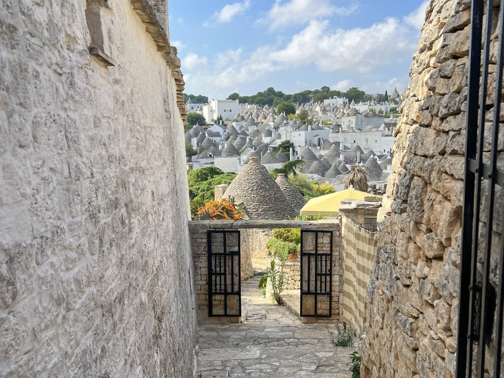 Rustic alleyway view overlooking the picturesque trulli houses in Alberobello, Puglia, Italy, showcasing charming stone walls, traditional architecture, and vibrant nature accents.