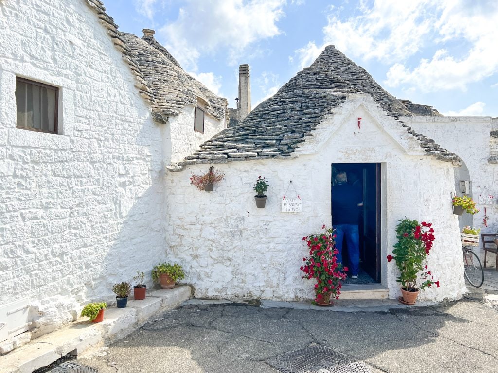 Charming white Trullo house in Puglia, Italy with traditional stone roof, colourful potted plants, and a visitor exploring the historic site, showcasing authentic architecture and southern Italian countryside charm.