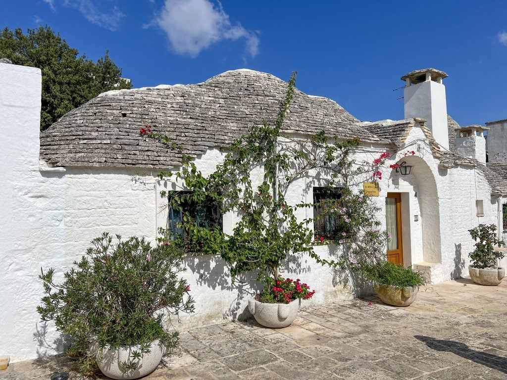 Charming white-trimmed trulli house with a conical roof in Alberobello, Italy, surrounded by colourful potted plants and vibrant bougainvillaea under a bright blue sky.