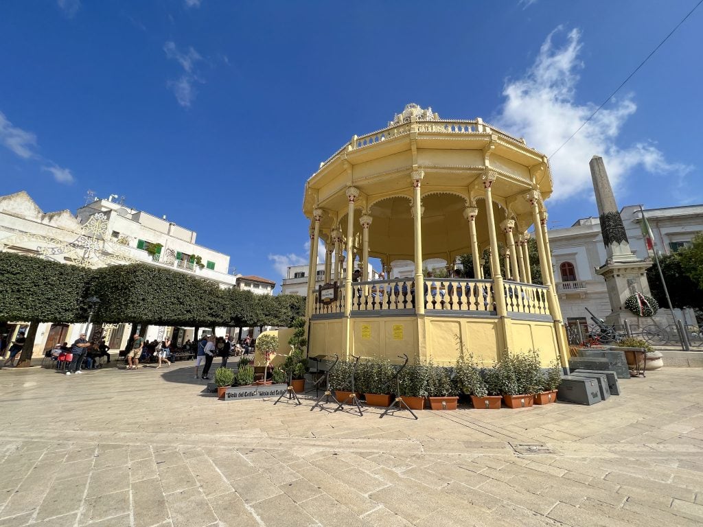 Victorian-era bandstand in a lively Italian piazza under a clear blue sky, surrounded by historic buildings, greenery, and tourists enjoying the sunny day.