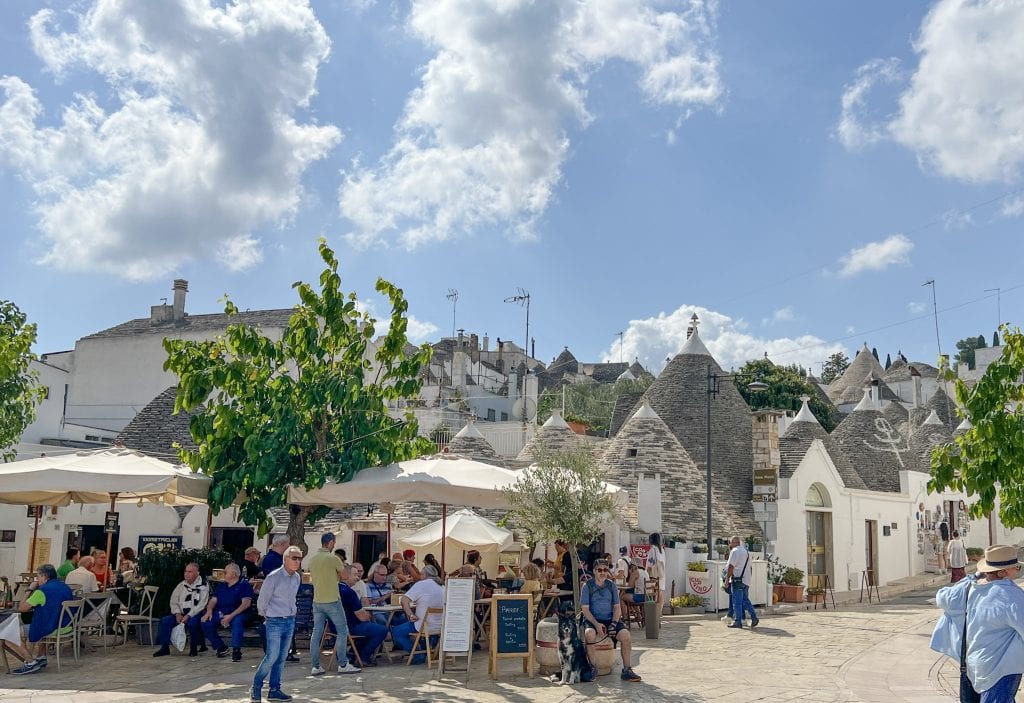 Vibrant outdoor café scene in Alberobello, Italy, with tourists enjoying sunny weather among iconic trulli houses under a bright blue sky.