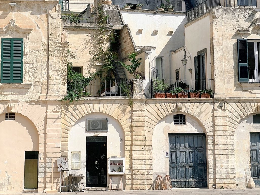 Charming historic limestone building with a shop on the ground floor and a balcony garden above, featuring vibrant plants and rustic shutters, embodying Mediterranean architectural charm.