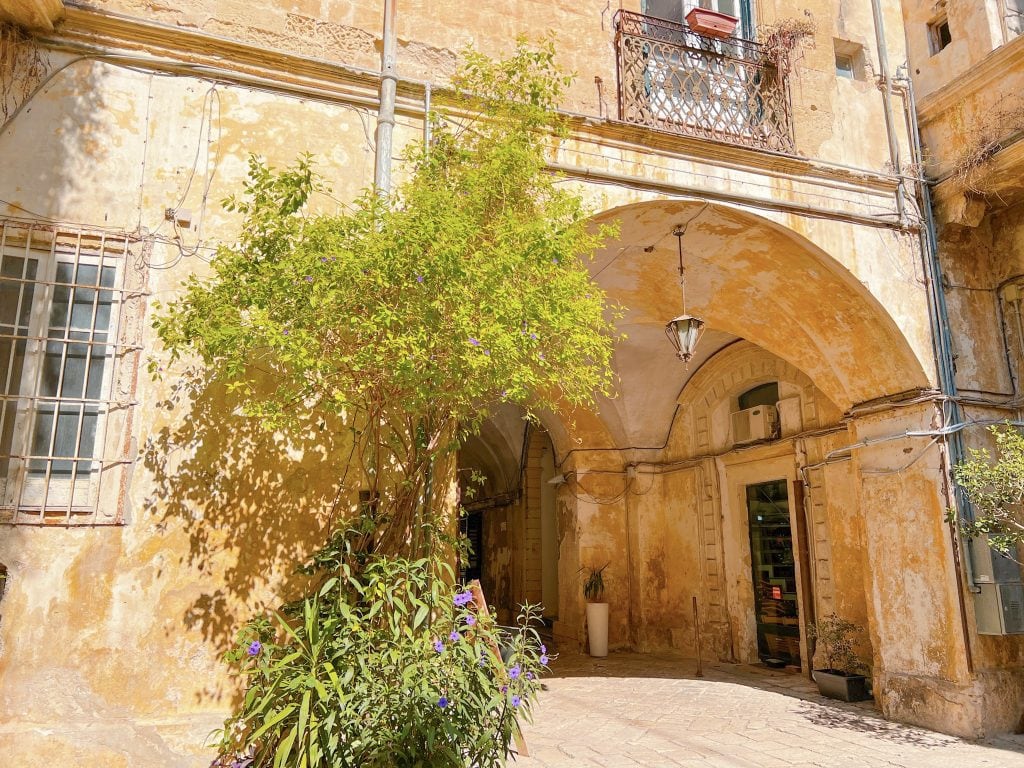 Vibrant alleyway with aged ochre buildings, lush greenery, and arched stone entrances in a charming Mediterranean village. Perfect for travel and exploration in Southern Europe.
