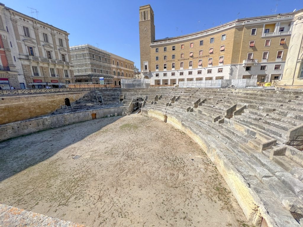 Ancient Roman amphitheatre in Rome, Italy, surrounded by historic buildings and modern architecture under a clear blue sky, showcasing Italy's rich cultural heritage and archaeological significance.